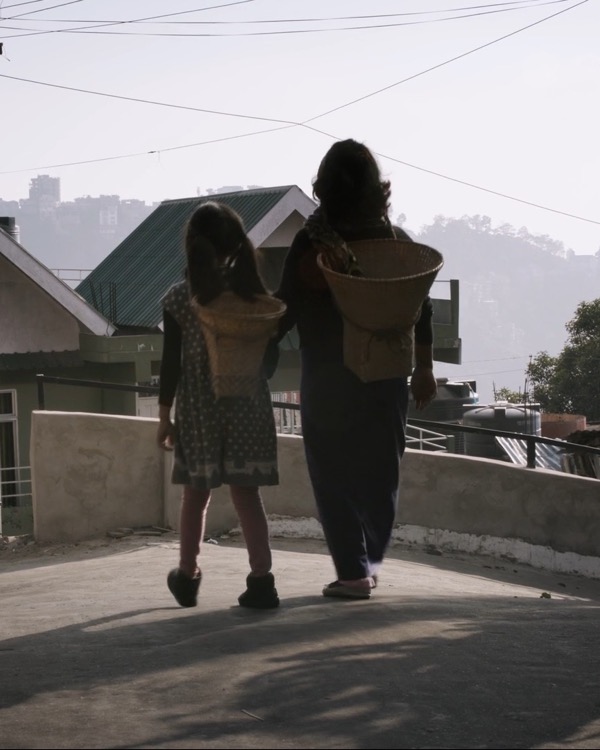 A frame from A Handful of Rice: a mother and daughter seen from behind, woven baskets on their backs, walking on above a misty valley